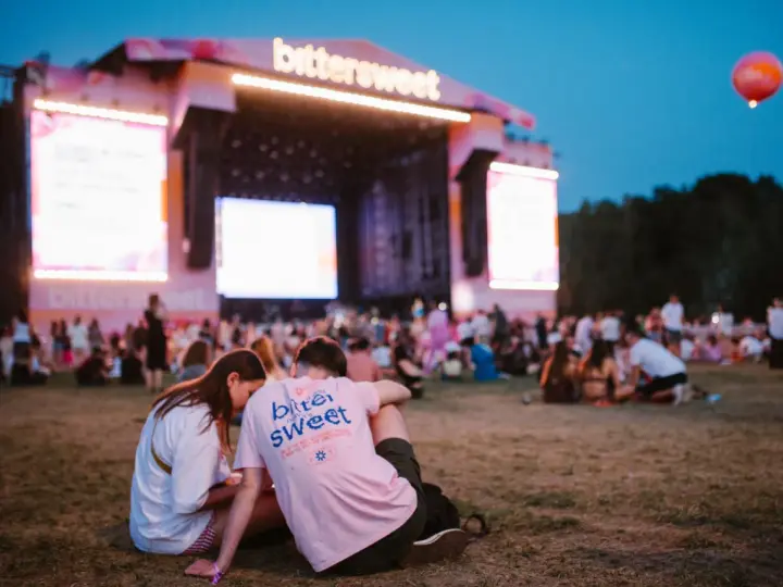 Para siedząca na trawie przed sceną festiwalową z napisem "bittersweet". W tle tłum ludzi i wielkie ekrany.