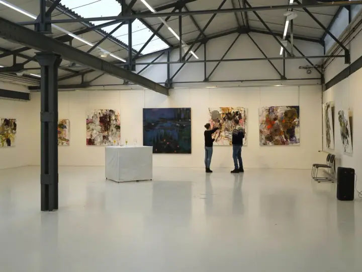 Zwei Personen betrachten Kunstwerke in der Kunsthalle Burkamp. Abstrakte Gemälde hängen an den weißen Wänden, ein Tisch mit Gläsern steht in der Mitte.