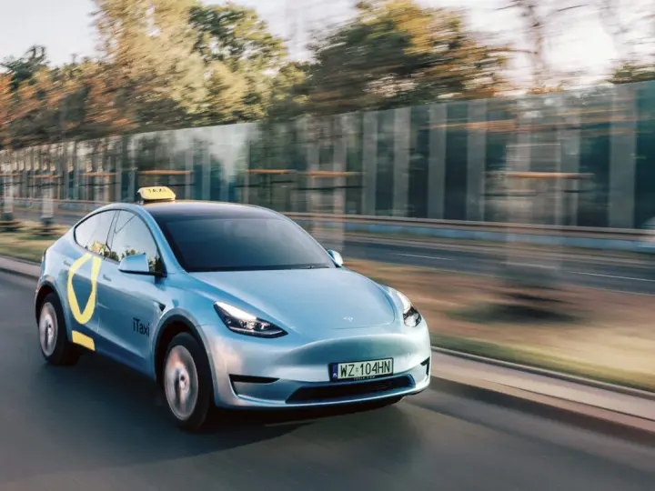 Niebieski Tesla Model Y z żółtym znakiem taxi na dachu i logo iTaxi na drzwiach, spełniający wymagania samochodu dla tej usługi.