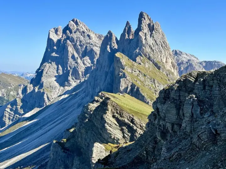Majestätische Felszinnen ragen in den blauen Himmel. Eine **Seceda Wanderung** führt durch grüne Almwiesen und felsiges Gelände.