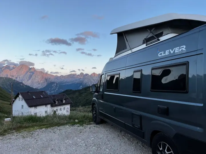 En mörkgrå husbil med pop-up tak, märkt "Clever vans", står parkerad vid en bergig stuga med Alperna i bakgrunden.
