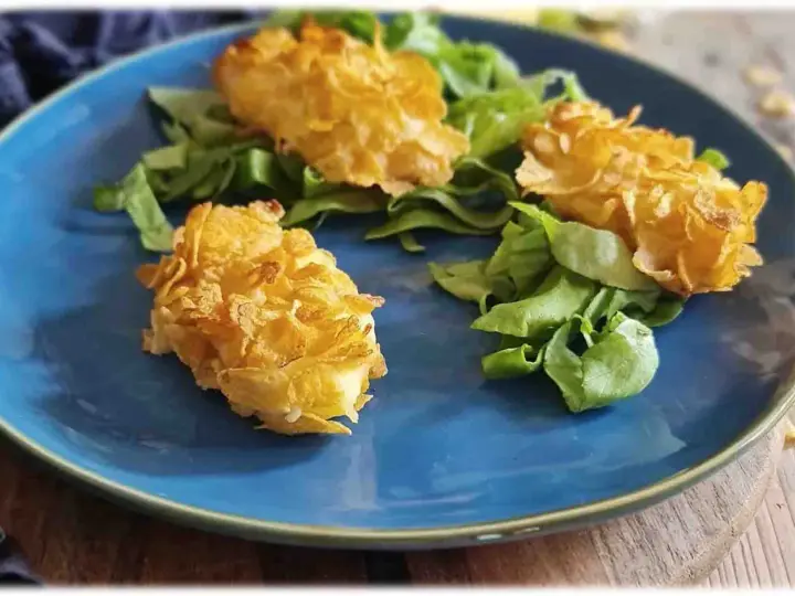 Tre croccanti polpette di patate impanate con fiocchi di mais, servite su un letto di insalata fresca su un piatto blu.