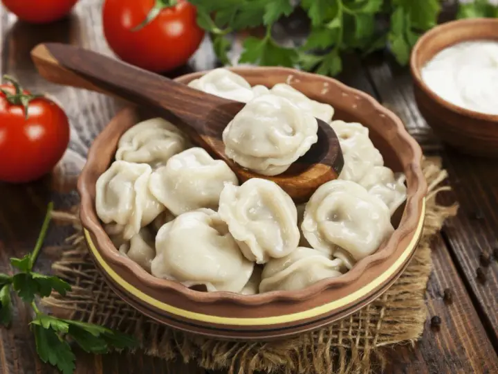 Pielmieni co to? Pyszne, gotowane pierożki w glinianej misce, z drewnianą łyżką, pomidorami i śmietaną w tle.