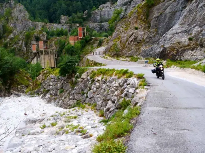 Un motociclista percorre una strada tortuosa tra le Alpi Apuane, con un fiume di sassi e ruderi industriali sullo sfondo.