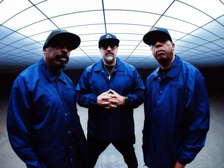 Les membres de Cypress Hill, B-Real, Sen Dog et DJ Muggs, posent ensemble, portant des vestes bleues assorties sous un plafond géométrique.