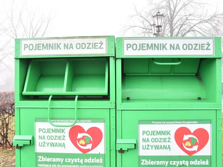 Dwa zielone kontenery na odzież używaną, z napisem "POJEMNIK NA ODZIEŻ UŻYWANĄ" i sercem z symbolami recyklingu.