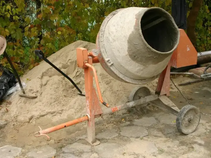Pomarańczowa betoniarka 250l gotowa do pracy, obok sterta piasku i łopaty.