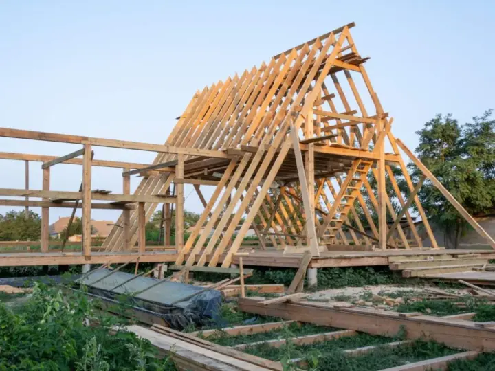 Drewniana konstrukcja domu w budowie. Widoczny szkielet dachu i ścian, schody. Fundament punktowy pod domek drewniany jest już gotowy.