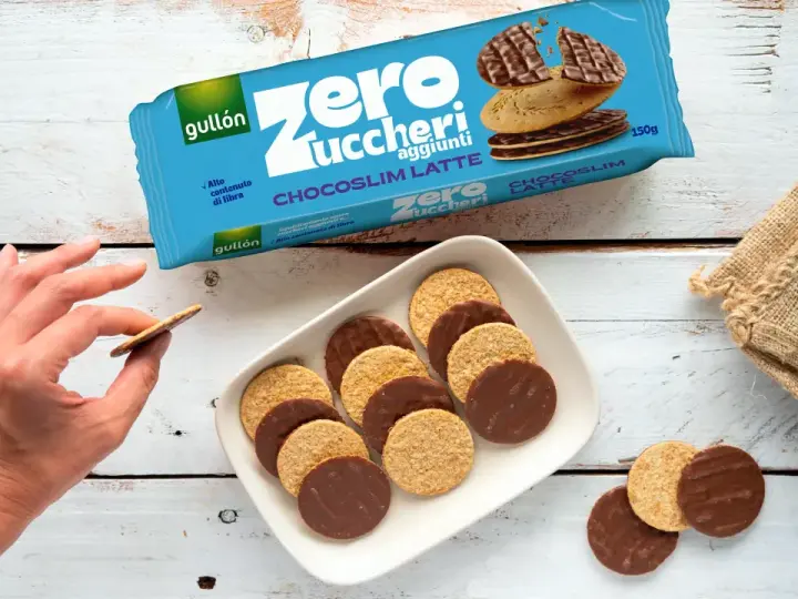 Biscotti senza zucchero Gullón Chocoslim Latte, alcuni ricoperti di cioccolato, su un piatto bianco. Una mano ne prende uno.