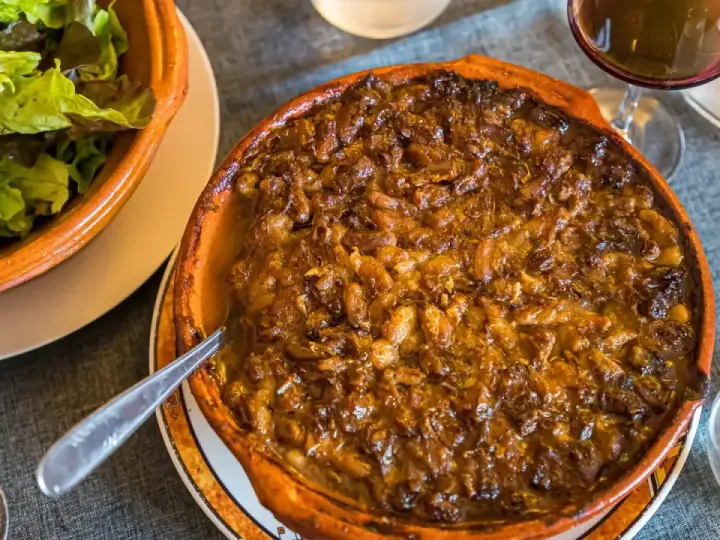 Cassoulet de Castelnaudary burbujeante en cazuela de barro, acompañado de ensalada verde y vino tinto.