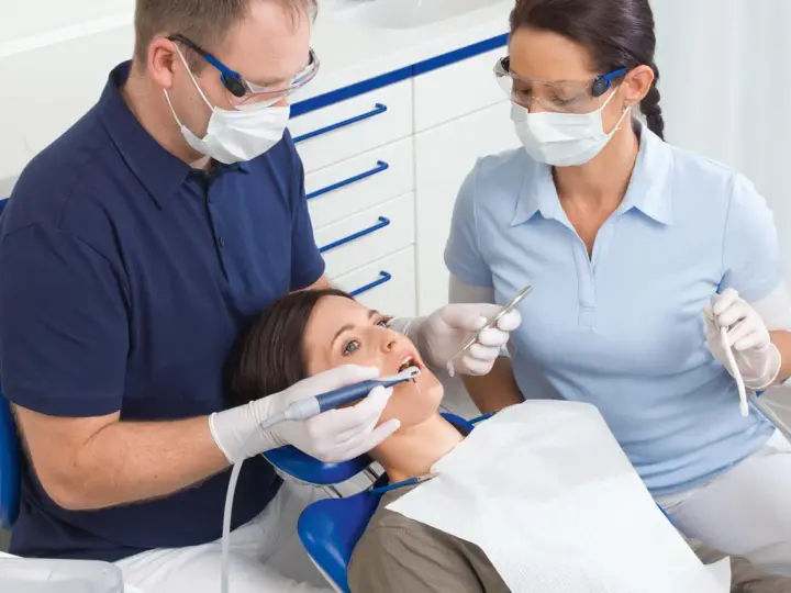 Dentysta i asystentka w okularach i maseczkach pracują przy pacjentce. Asystentka trzyma narzędzie, a dentysta używa **vector paro** do zabiegu.