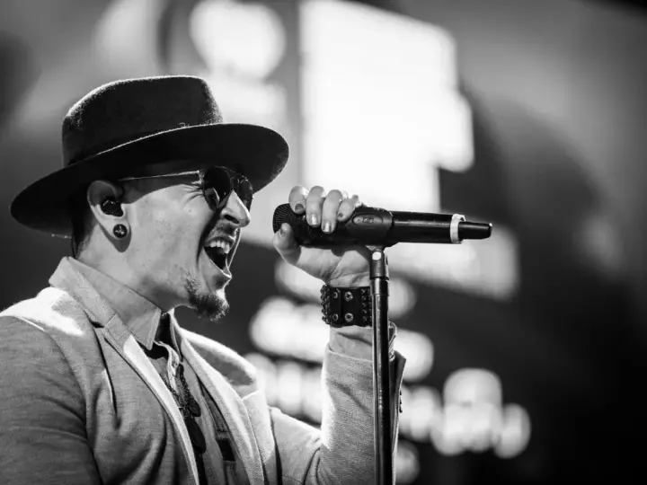 Wokalista Linkin Park, Chester Bennington, w kapeluszu i okularach przeciwsłonecznych, śpiewa do mikrofonu z pasją.
