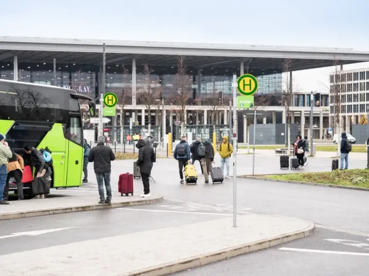 Ludzie z bagażami wsiadają do zielonego autokaru przed nowoczesnym budynkiem, przypominającym lotniska w Berlinie.