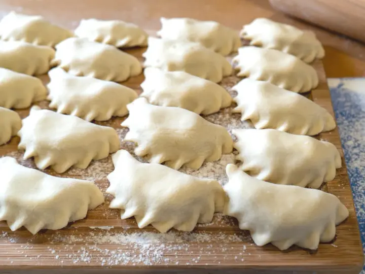 Surowe pierogi ułożone rzędami na drewnianej desce, posypanej mąką.