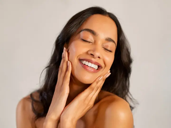 Une femme au sourire radieux caresse son visage, révélant une peau glowy éclatante de santé.