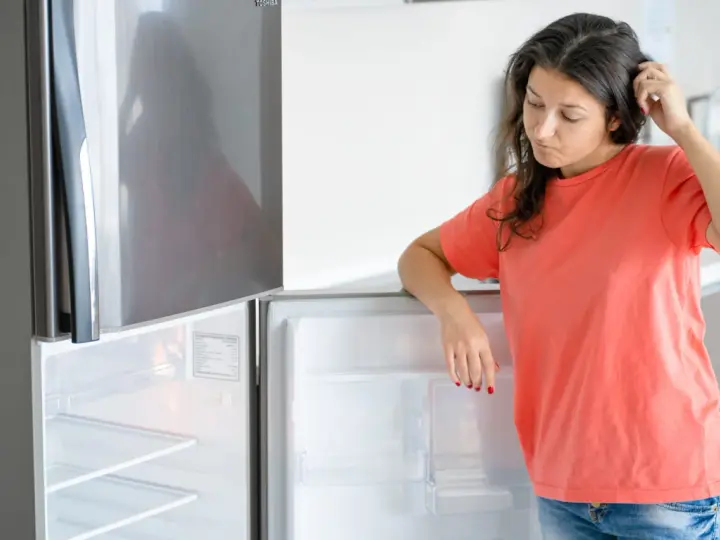 Une femme perplexe regarde son frigo vide, qui fait un bruit étrange. Elle se gratte la tête, se demandant ce qu'elle va manger.