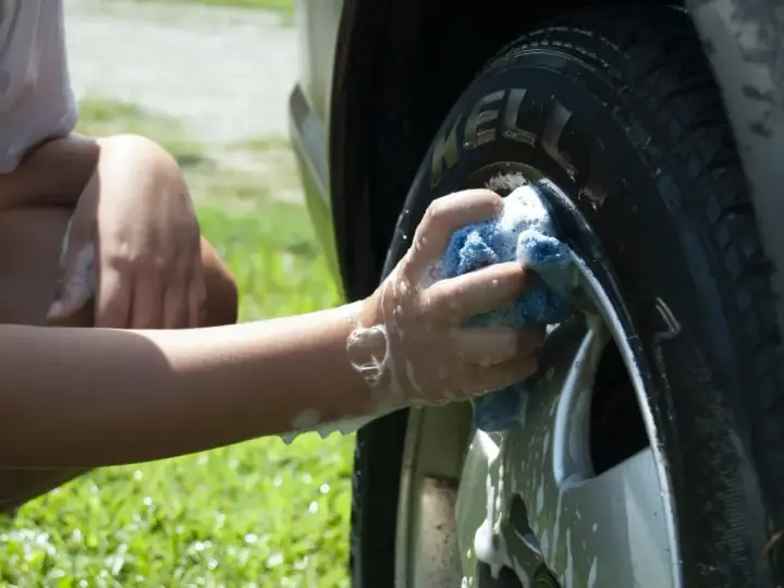 A person's arm and hand, covered in suds, scrub a car tire with a blue sponge. It seems they are testing if you can use dish soap to wash your car.