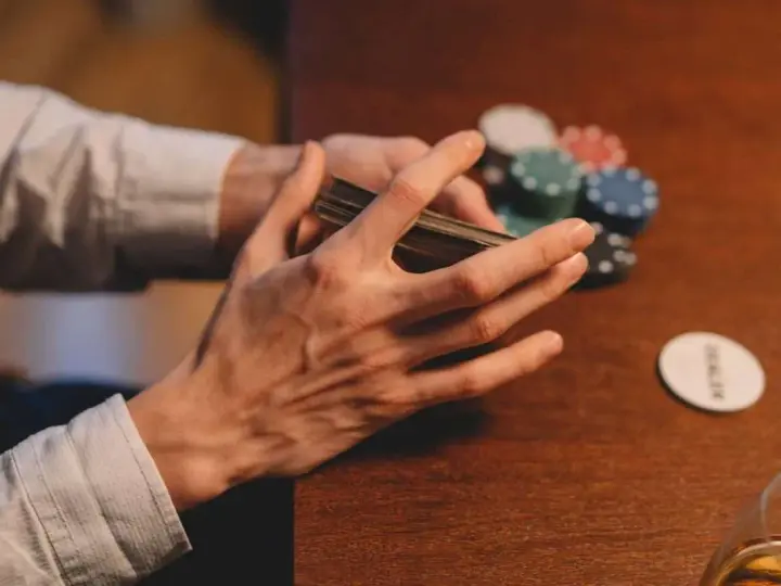 Händer som blandar kort vid ett pokerbord, med marker och en "dealer"-bricka. En påminnelse om att spelberoende behandling finns tillgänglig.