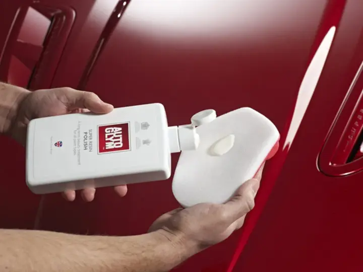 Händer applicerar Autoglym Super Resin Polish på en blank röd bil med en svamp.