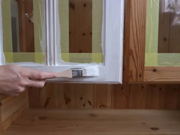 En hand målar en träyta vit med en pensel. Maskeringstejp skyddar intilliggande ytor av furu.