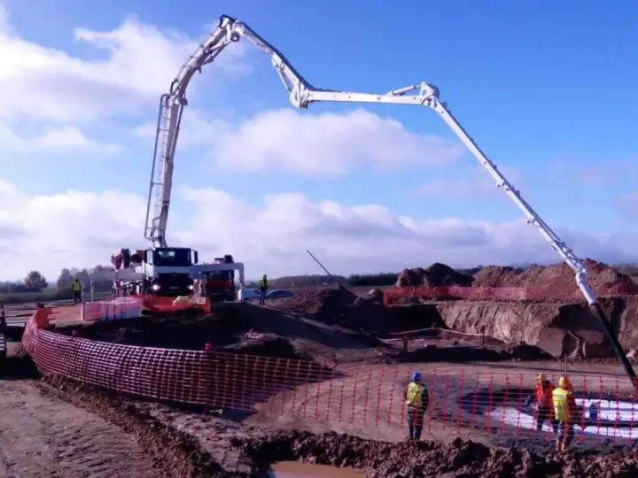 Ogromna pompa do betonu pracuje na budowie, dostarczając materiał do wykopu. Pracownicy w kamizelkach obserwują proces.