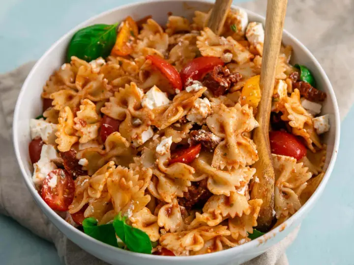 Ein köstlicher, sommerlicher Nudelsalat mit Farfalle, Tomaten, Feta und Basilikum, serviert in einer weißen Schüssel mit Holzutensilien.