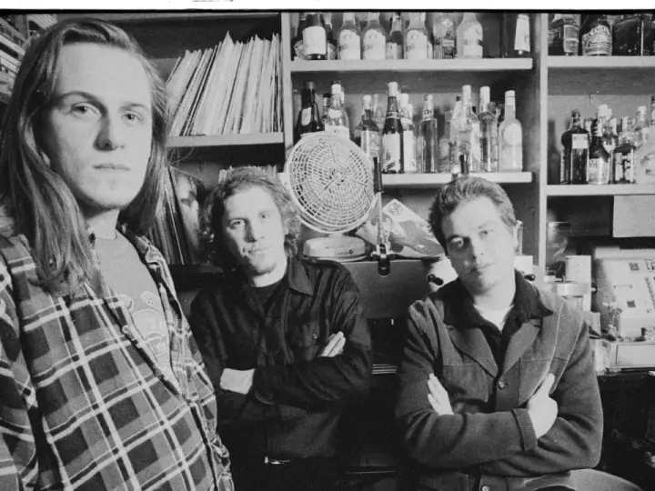 Tres hombres posan en blanco y negro, con estanterías de discos y botellas detrás.