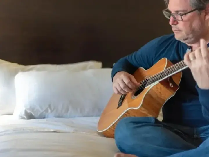 Mężczyzna w okularach gra na gitarze siedząc na łóżku. Gdzie ta keja nuty?