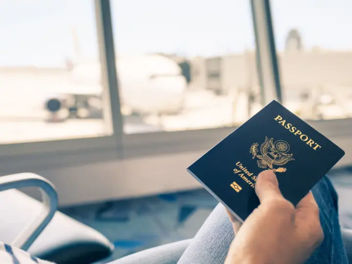 Main tenant une carte d'identité américaine devant une fenêtre d'aéroport avec un avion en arrière-plan.