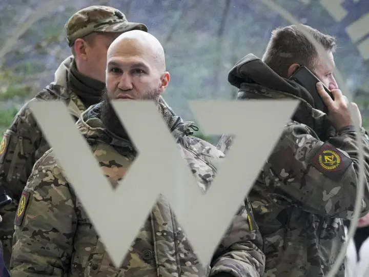 Trzech mężczyzn w mundurach, jeden rozmawia przez telefon, na tle symbolu przypominającego literę "W". Mogą to być członkowie grupy Wagnera.