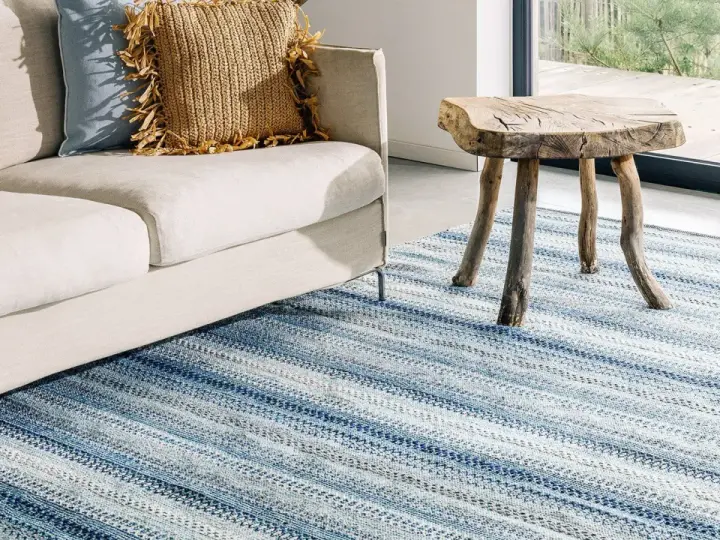 En ljus soffa med kuddar och en rustik pall står på en randig blå matta. Perfekt inspiration matta vardagsrum för en avslappnad stil.