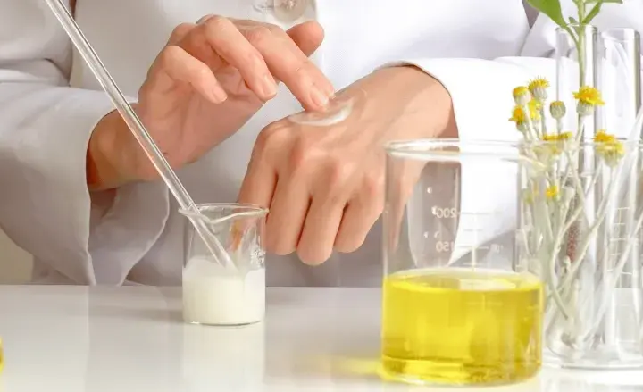 Manos de científico aplicando crema, con tubos de ensayo y líquidos amarillos. El cetearyl alcohol es clave en esta formulación.