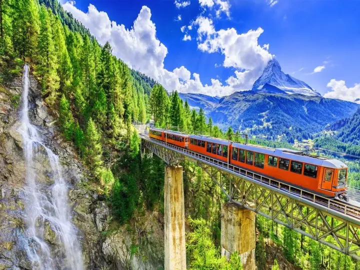 An orange train crosses a bridge over a waterfall, on its way from Zurich to Andermatt. Lush green trees and mountains surround the scenic route.