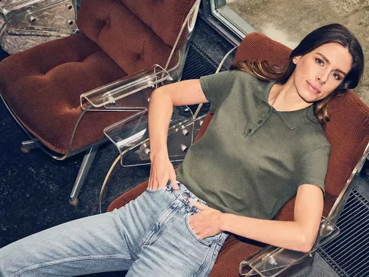 Mujer con vaqueros y polo verde, sentada en una silla de terciopelo, encarnando la moda atemporal.
