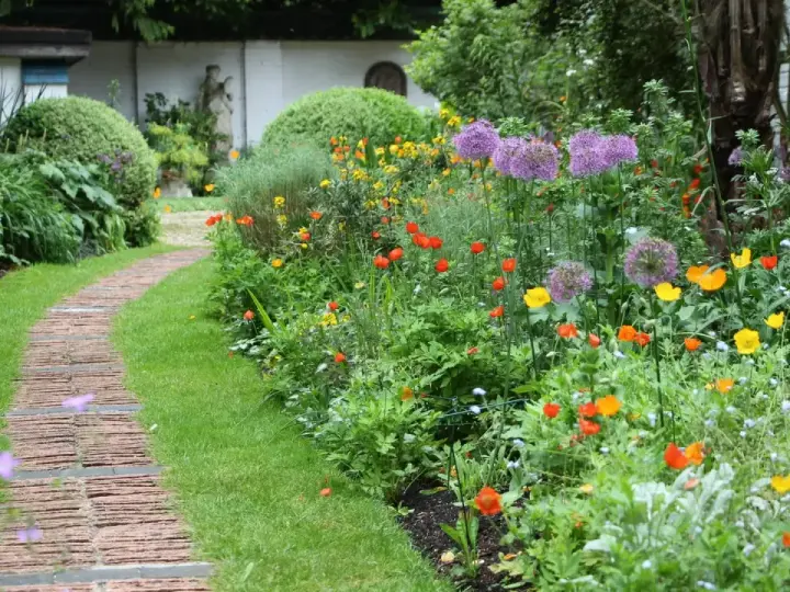Ein geschwungener Weg aus Ziegelsteinen schlängelt sich durch einen üppigen bauerngarten mit leuchtenden Blumen und grünem Rasen.
