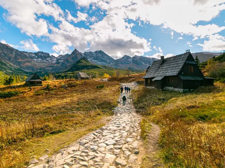 Kamienista ścieżka prowadzi do Rusinowej Polany, gdzie widać drewniane chaty i turystów. W tle majestatyczne Tatry.