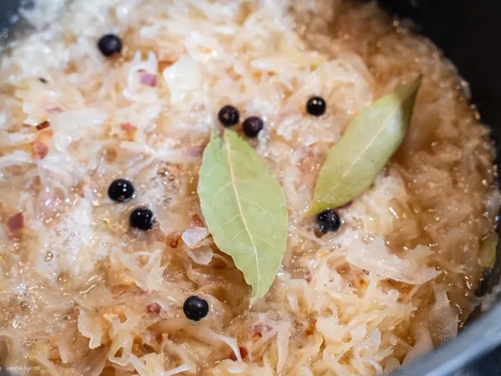 Sauerkraut köchelt mit Lorbeerblättern und Wacholderbeeren, um den Geschmack zu verfeinern. Kleine rote Zwiebelstücke sind ebenfalls sichtbar.