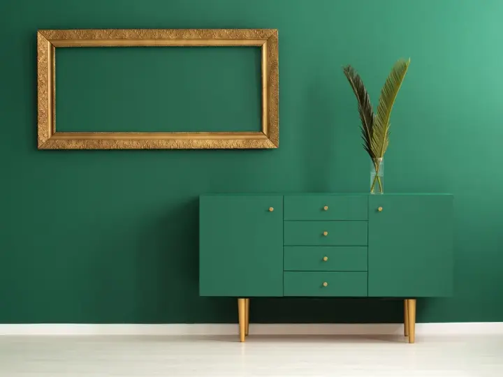 Un mueble verde esmeralda con patas doradas y un marco dorado vacío sobre pared verde. Los colores que pegan con el dorado son el verde intenso y el blanco.