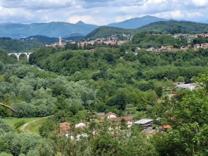 Panoramica collinare con un ponte ad archi, un campanile e case sparse a Cassano Magnago, circondata da rigogliosa vegetazione.