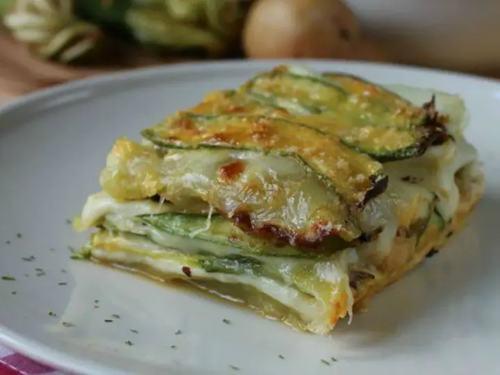 Una fetta di parmigiana di zucchine, stratificata con formaggio filante e zucchine grigliate, servita su un piatto bianco.