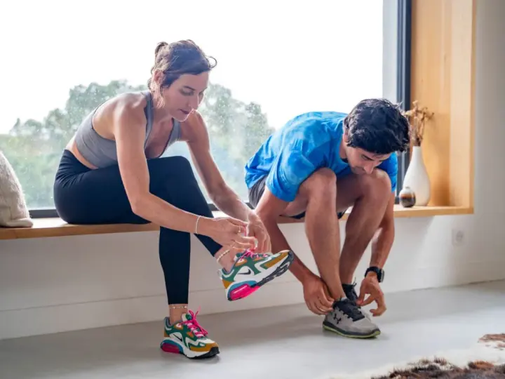 Un couple se prépare pour reprendre le sport, enfilant leurs chaussures de course près d'une fenêtre lumineuse.