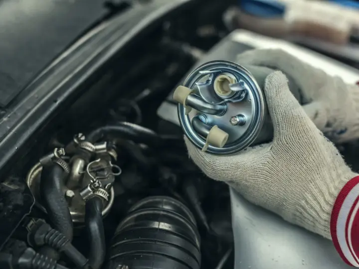 Hände mit Handschuhen halten einen neuen Kraftstofffilter bereit, um den alten im Motorraum zu wechseln.