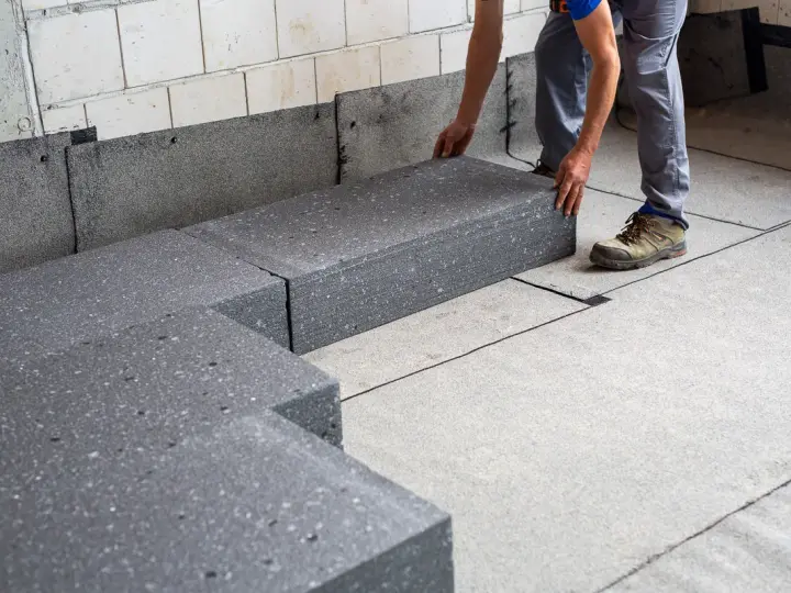 Pracownik układa szary styropian pod podłogówkę. Elementy tworzą izolację na betonowym podłożu.