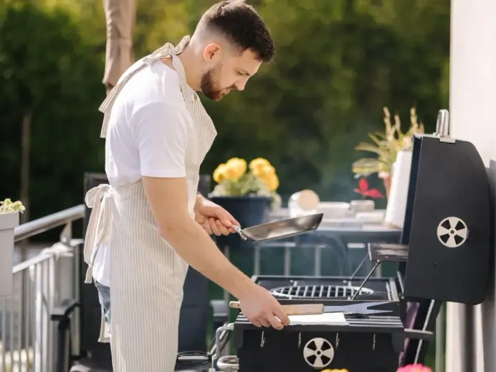 Mężczyzna w fartuchu przygotowuje grilla na balkonie, co pokazuje, że można robić grilla na balkonie.