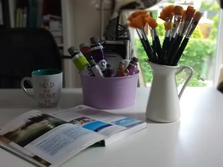 Pędzelki malarskie w dzbanku, tubki farb w fioletowym wiaderku, kubek z napisem i otwarta książka.