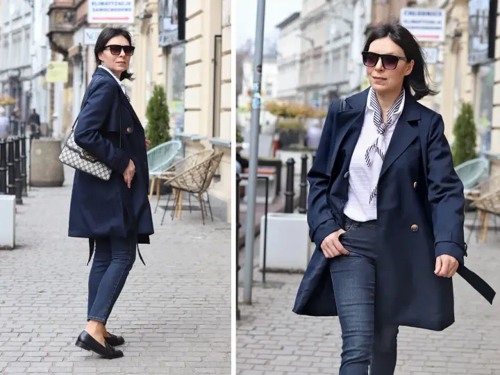 Elegancka kobieta w granatowym trenczu damskim, białej bluzce w paski i jeansach spaceruje po brukowanej ulicy.