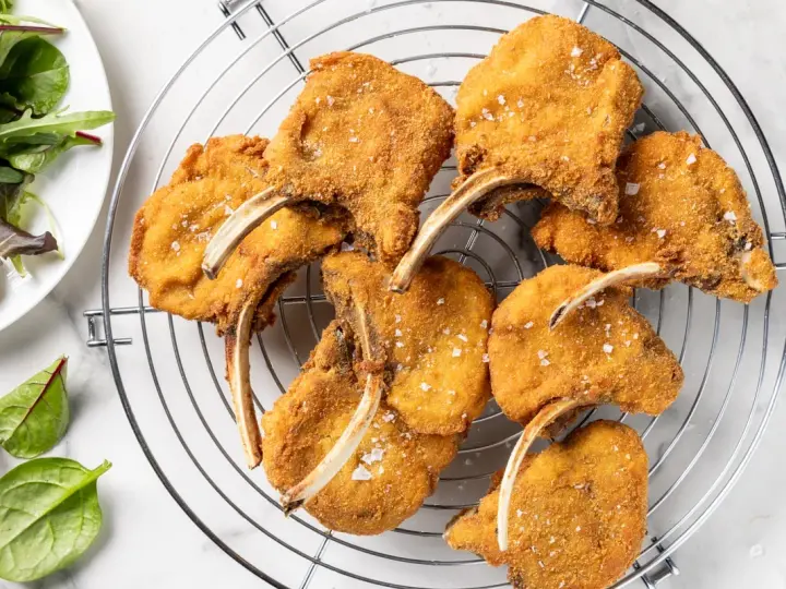 Deliziose costolette d'agnello fritte, dorate e croccanti, cosparse di sale grosso, su una griglia. Accanto, un'insalata fresca.