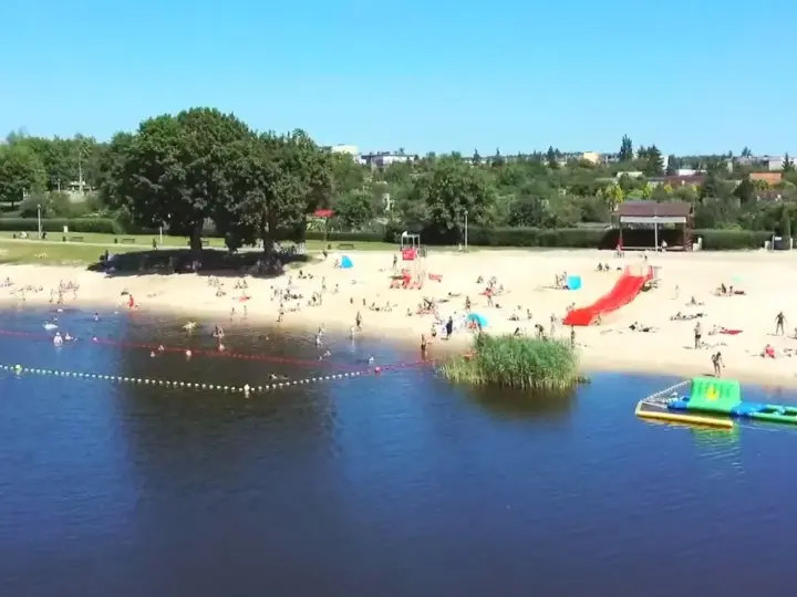 Słoneczny dzień nad jeziorem Grzymisławskim. Ludzie odpoczywają na plaży, kąpią się i bawią na dmuchanych atrakcjach.