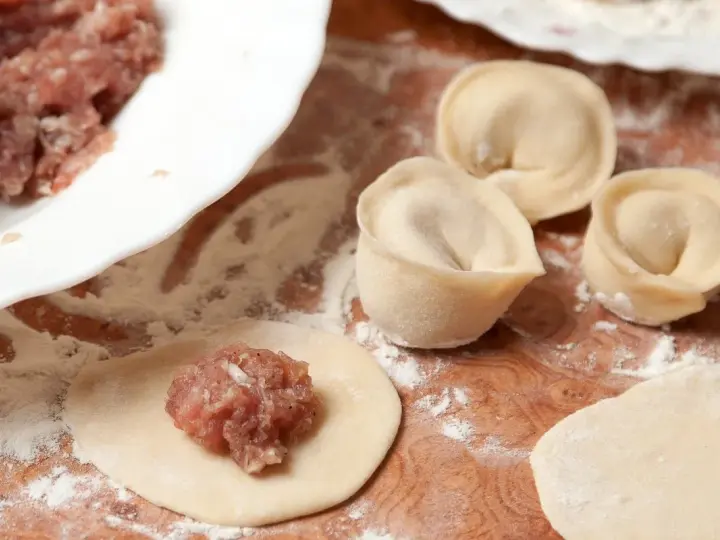 Przygotowanie pierogów z mięsem mielonym surowym. Na drewnianej desce widać surowe ciasto, farsz i gotowe pierożki.