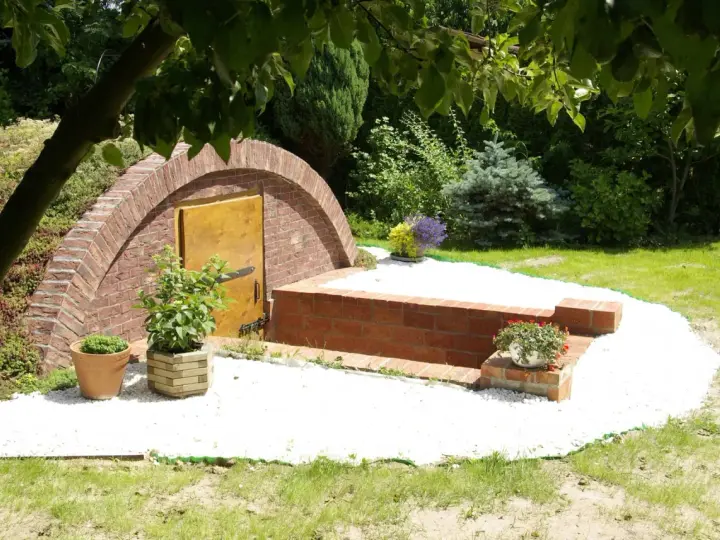 Ziemianka z cegły z drewnianymi drzwiami, otoczona białym żwirem i zielenią. Zastanawiasz się, ile kosztuje ziemianka?